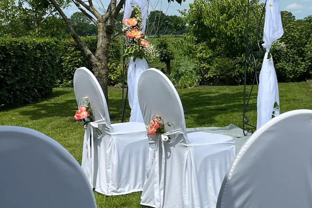 Zwei weiß bezogene Stühle mit rosa Blumenarrangements und weiß drapiertem Traubogen auf einer grünen Wiese