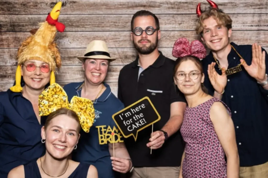 Fünf Personen posieren in einer Fotobox mit Party-Accessoires; Sprechblasen-Schild: I'm here for the CAKE! und Team Bride