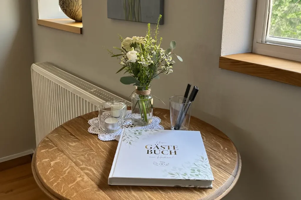 Gästebuch auf rundem Holztisch neben Vase mit Blumen, Kerzen und Stiften am Fenster